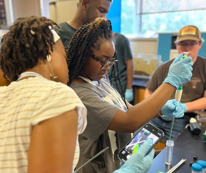 Students process water samples in a laboratory. One student holds an instrument in a tube of water while several other students look on. 