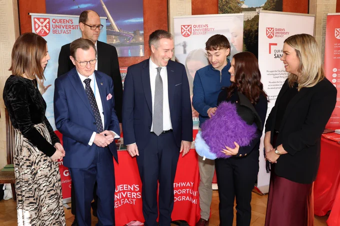 Image shows Paul Givan, Education Minister, pictured with Professor Sir Ian Greer, Queen's staff members, and Professor Fluffy. They are pictured in front of a promotional stand for our Pathway Opportunity programme, with pop-up stands and a red branded tablecloth.