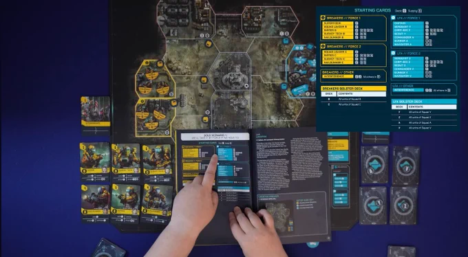 A screengrab from a Beyond Solitaire YouTube video showing setup for the solo mode of the board game Undaunted 2200: Callisto, with a pair of hands pointing to the rulebook and a game board laid out with decks of cards and various cardboard tokens.