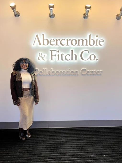 Elleanor Ross standing in front of an Abercrombie & Fitch Co. Collaboration Center” sign. The sign is all white with the letters glowing. She is wearing a white turtleneck, brown jacket, a beige skirt with a silver hoop belt, and brown boots. Photo courtesy of Elleanor Ross.