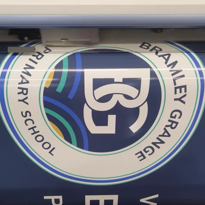 A large format printer producing a graphic for a sign at Bramley Grange Primary School.  The graphic features a circular logo for the school with a 'B' and 'G' interlocked with the text 'Bramley Grange Primary School'.  