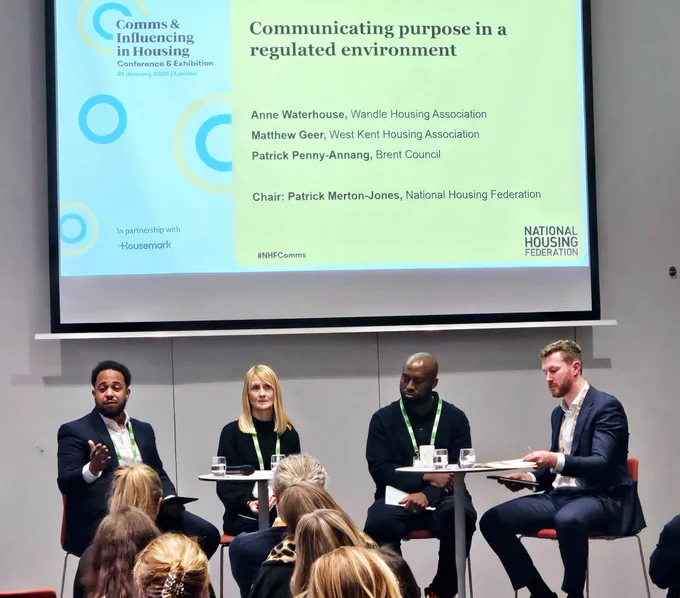 A photo of four people sitting on stools at a conference talking to an audience.