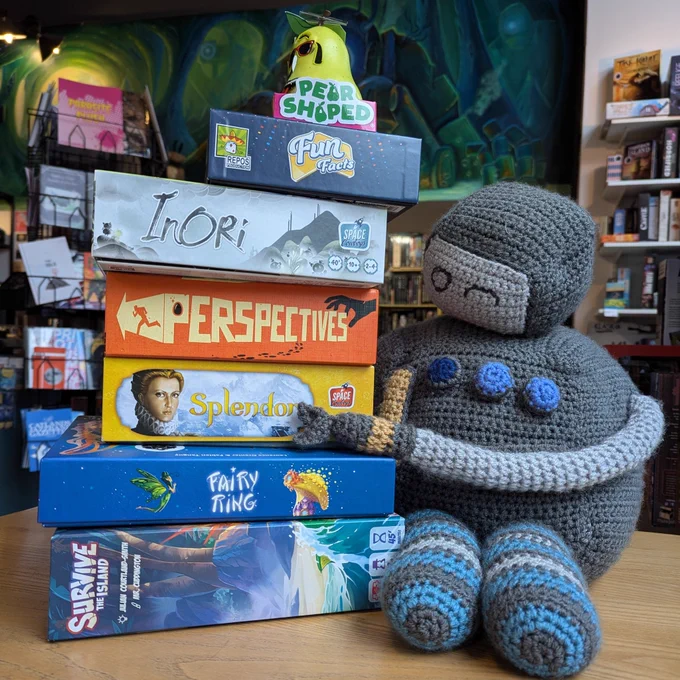 A crocheted robot holding onto a stack of board games in the Edinburgh board game shop Ancient Robot Games. The board games include Pear Shaped, Fun facts, Inori, Perspectives, Splendor, Fairy Ring and Survive the Island.
