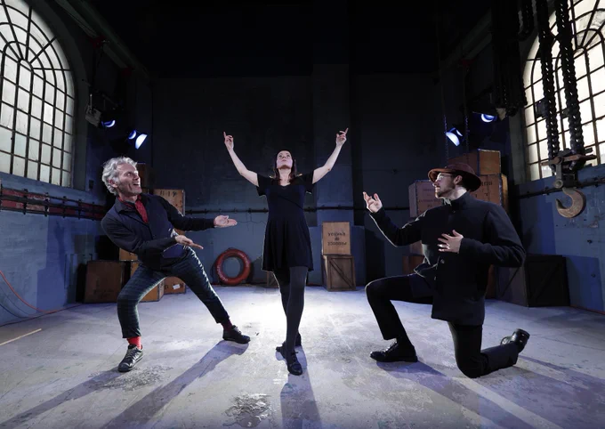 Three performers are caught in motion in the centre of a large warehouse space. Two men on bended knees point to a central woman who raises her arms to the ceiling.