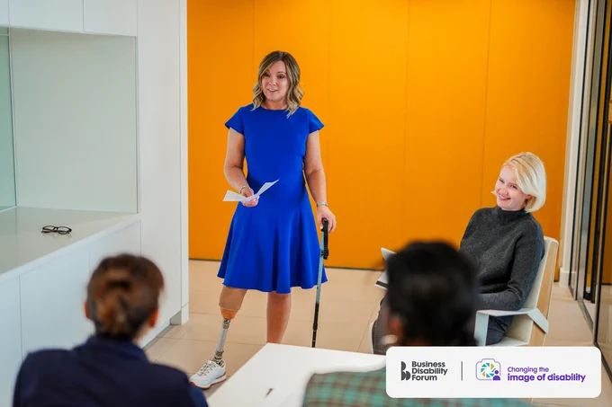 Professional in a blue dress, who wears a prosthetic leg and uses a stick, stands in front of 3 colleagues during a meeting. The Business Disability Forum 'Changing the image of disability' logo is in the lower right corner of the image.
