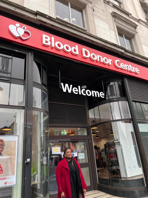 Bell stands outside a Blood Donor Centre with a red facade and the Give Blood logo on it. There is a big sign that says 'Welcome' above her head. She wears a red coat.