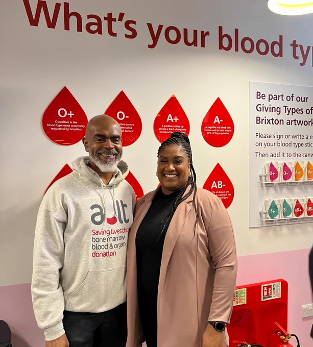 Bell stands next to man wearing a grey ACLT hoodie in front of a wall display that says 'What's your blood type?' and lists the various blood groups.