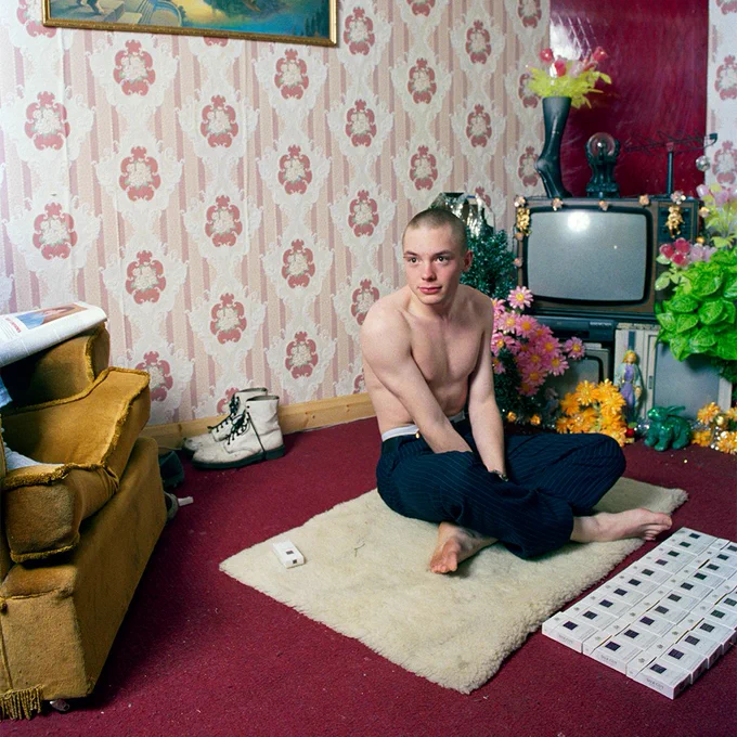 Jamie Lloyd sits on a bold pink carpet with their legs crossed in the middle of a room, it appears to be early morning as the light indicates so. They are topless and have no shoes or socks on, just a pair of grey trousers. One might assume they have been partying all night and just got home! There are lots of packets of cigarettes laid out neatly in front of them. There are lots of other items in the room, some dr marten boots in white behind them, a sofa chair, a TV, other living room clutter. The room is decorated with bright colours. 