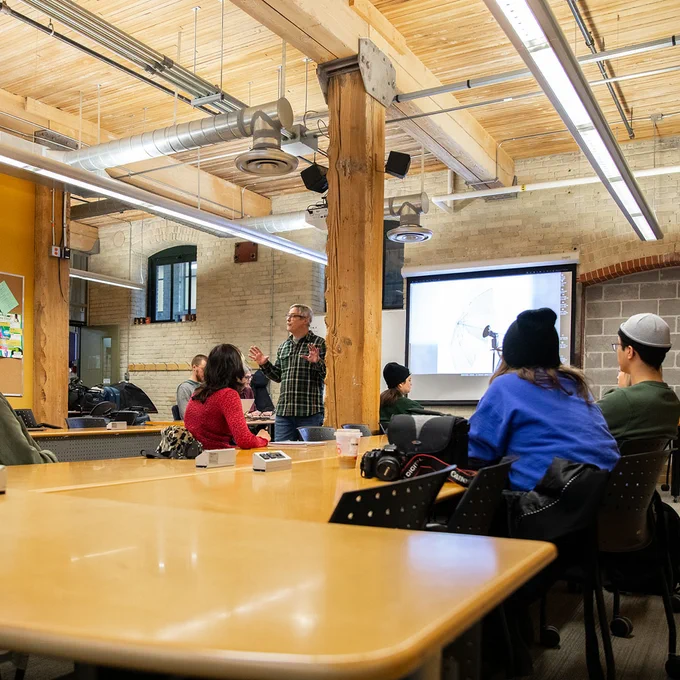 Students attentively listening to an Instructor during a Creative Communications class in a modern, industrial-style learning space.