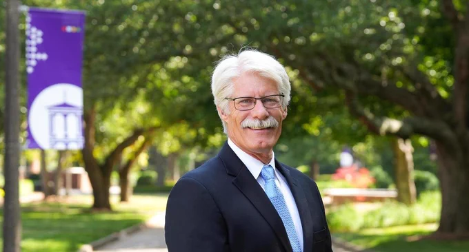 A white man with white hair wears glasses and dark suit outdoors. A purple college banner appears in the background.