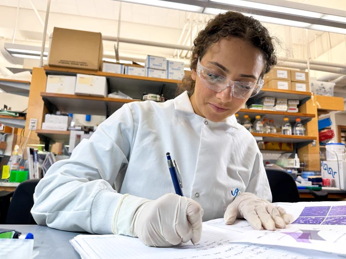 A JAX Trainee, wearing a white lab coat, gloves and safety glasses, writes in a lab notebook at a lab bench.
