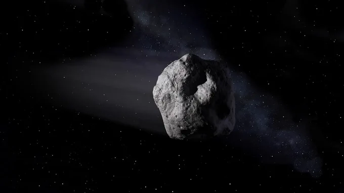 An artist's concept of an asteroid floating in space. The asteroid appears rugged and irregularly shaped, with visible craters and textured surface details. A faint trail of dust or gas extends from the asteroid, set against a backdrop of a star-filled sky and distant nebulous clouds.