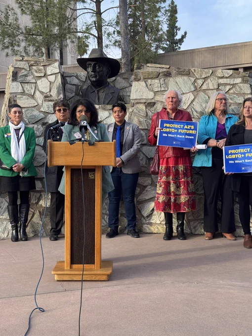 Democratic Rep. Betty Villegas speaks into microphones perched on a wooden podium in the Rose Garden outside of the Arizona State Capitol. Supporters, including Democratic Reps. Patty Contreras and Lorena Austin, stand behind Villegas. Two people on the right hold blue Human Rights Campaign signs that read “Protect AZ’s LGBTQ  Future! We Won’t Back Down.”