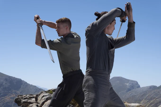 Rand al'Thor (Josha Stradowski) and Lan Mandragoran (Daniel Henney) wield swords back to back against a blue skied mountainscape.