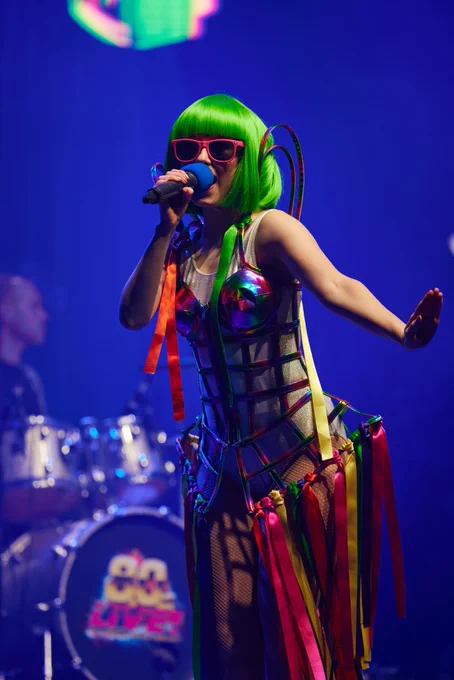 A woman on stage singing for 80s Live with neon green air, pink glasses and a blue microphone