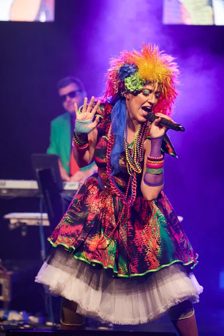 A woman on stage dressed in 80s clothing with big colourful hair, lots of neon beads and fingerless gloves. 