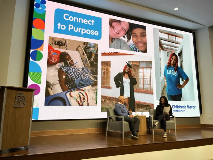 A presentation at Children's Mercy Kansas City, featuring a large screen displaying the message "Connect to Purpose" alongside photos of various individuals engaged in different activities. In the foreground, three people are seated on stage, engaged in conversation next to a lectern with the Children's Mercy logo.