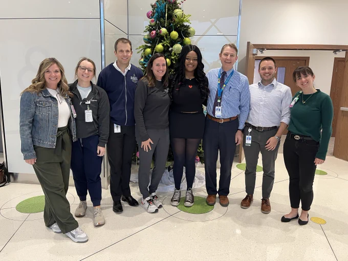 Group of eight professionals posing together in an office, smiling in front of a decorated Christmas tree.