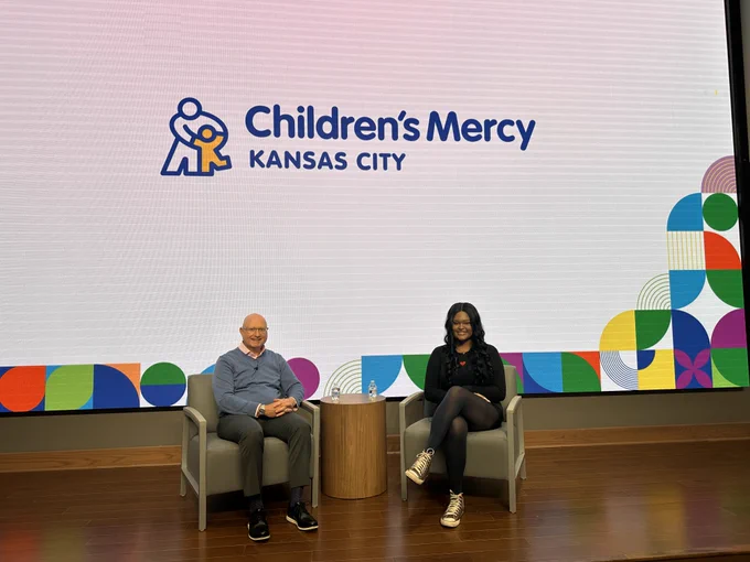 Two individuals seated on stage chairs in front of a presentation screen displaying the logo of Children's Mercy Kansas City.
