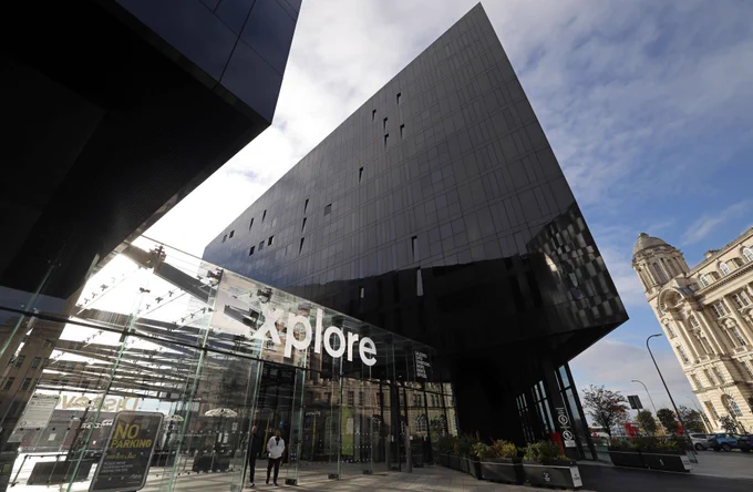 The modern building on Mann Island which has a striking angular design and features the word 'Explore' prominently above the glass entrance, under a clear blue sky. Two people are standing by the entrance.