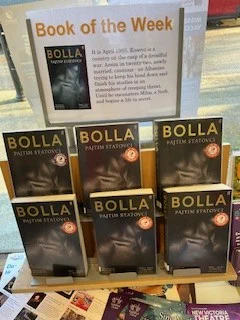 Image is a photograph of Horsley Library's Book of the Week display, a plastic stand featuring 6 copies of Pajtim Statovci's "Bolla". Above the display is a laminated advertisement in landscape format. Said advertisement is titled: 'Book of the Week' and has a picture of the Book's cover on the left and the text of the blurb on the right, as in the body of the post.