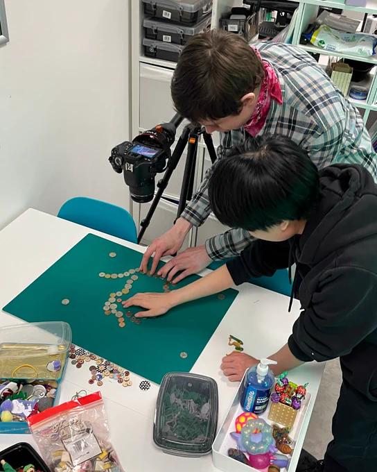 Two young participants work together on a stop-motion animation project, arranging coins into a creative pattern on a green board. A camera on a tripod is set up to capture their work, surrounded by various craft supplies.