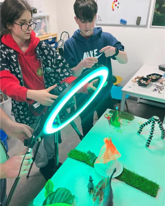 Two participants collaborate on a stop-motion animation scene involving a toy dinosaur and props like a teepee made from skewers and orange fabric. A ring light, illuminating a green light over the scene, and camera are set up to capture the action.