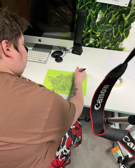 Close-up of a young person drawing intricate black ink designs on a bright green piece of paper. Their arm, with a tattoo of flowers, is visible as they focus on their work.