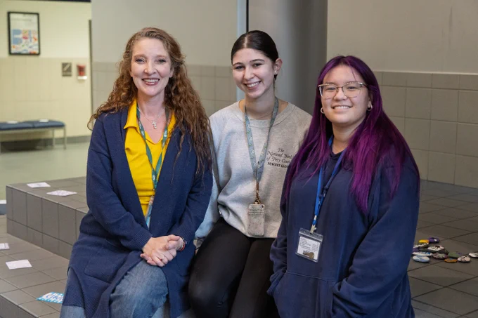 Three women pose for a photograph; all three are casually dressed, smiling, and wearing name badges.