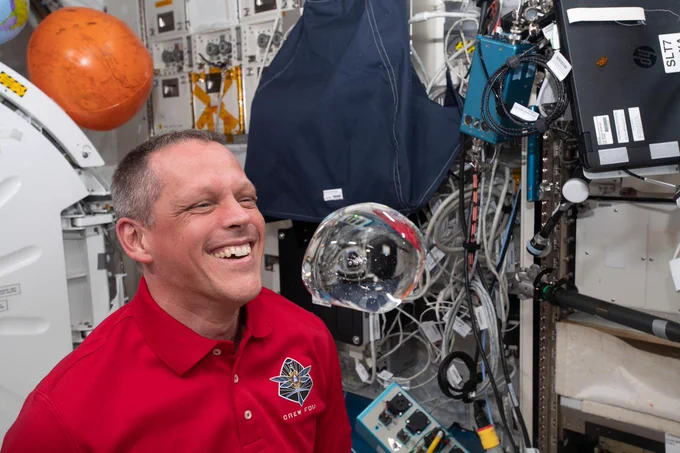 Robert Hines, wearing a red polo shirt with his mission logo on the front, smiles widely as he looks at a floating water bubble aboard the International Space Station.