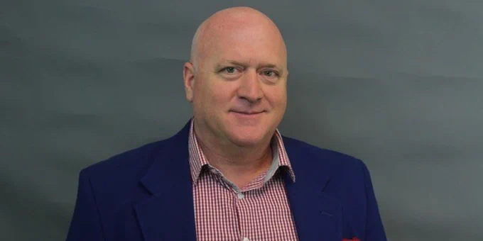 Portrait of Clint Ballinger, smiling in a blue blazer and red checked shirt against a grey background.