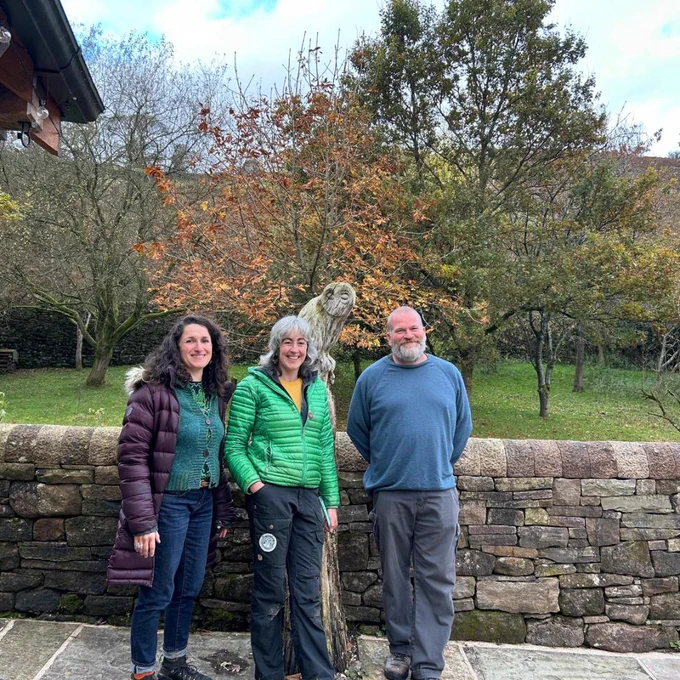 3 people stood outside in front of a stone wall smiling.