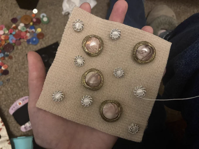 A square cream coloured knit cloth on a hand. On the cloth there are 12 jewel-like decorative buttons sewn. In the background, there are various sewing equipments on floor such as thread and buttons.