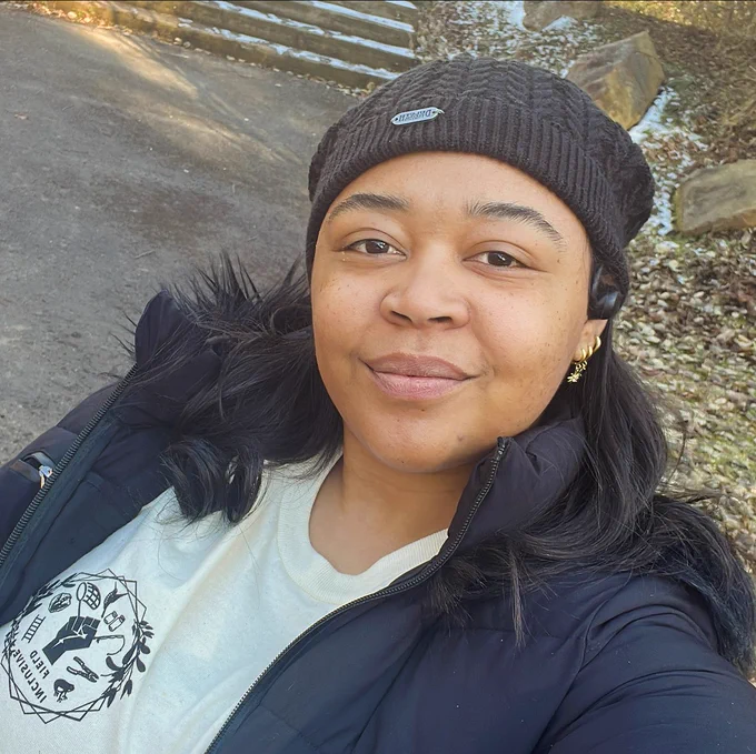 Lauren taking a selfie wearing a black coat, black beanie, and beige tshirt with a logo.