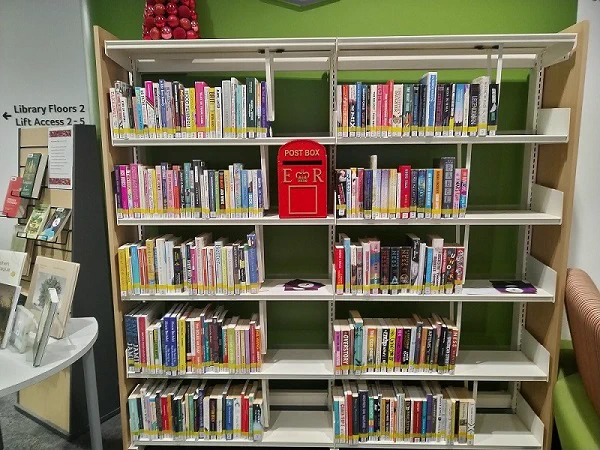Interior of a library showing well-organized bookshelves filled with books. In the center, a red post box labelled "Eco R" is featured for book returns. Adjacent seating area and library signage visible.