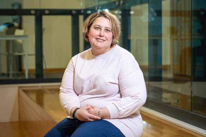 An individual seated in an indoor setting, wearing a pink long-sleeve top and blue pants, smiling at the camera. The background features a glass barrier and reflections on a polished wooden floor.