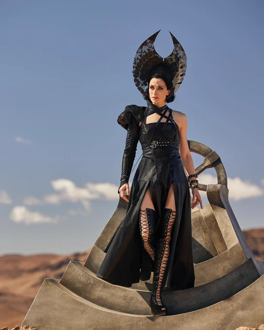 Natasha O’Keeffe as Lanfear in The Wheel of Time Season 2, standing on a stone platform in an elaborate black outfit with a striking crescent-shaped headdress, set against a clear blue sky and a rocky desert landscape