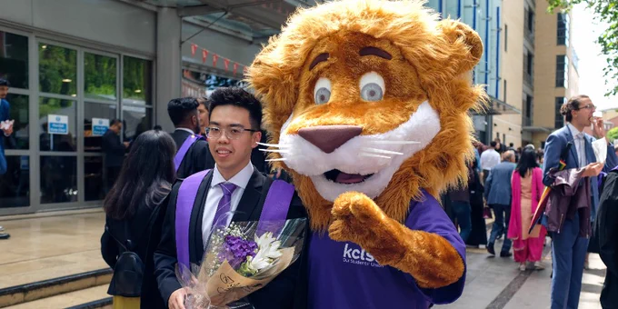 A graduate wearing a gown with purple accents, smiles while holding a bouquet of flowers. Beside him stands cheerful lion mascot.