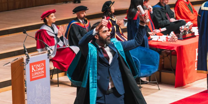 A graduate in formal attire and academic robes celebrates energetically on stage, holding up their diploma. 