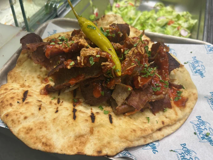 A serving of kebab on a naan bread topped with sliced meat, sauce, and a hot pepper, presented on a tray with a printed logo.
