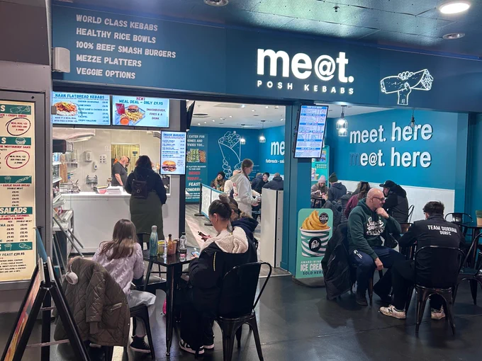 Interior of a busy food court with customers seated and ordering at the MEAT Posh Kebabs stand, offering kebabs, burgers, and salads.