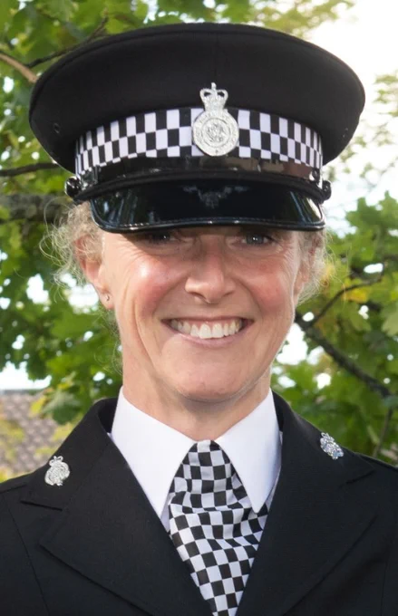 A picture of a smiling female police officer in uniform. She wears a tunic, chequered neck tie and flat hat. 