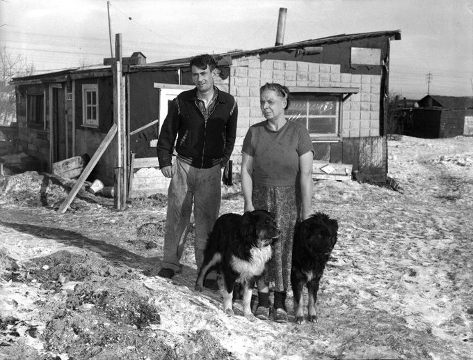 This black-and-white photo depicts a man and a woman standing outside a modest, possibly makeshift home in a rural or remote area. The house appears to be constructed from a combination of wood and other materials, with visible signs of wear and makeshift repairs. The ground is covered with patches of snow, indicating a cold climate. Both individuals are dressed in simple, practical clothing suitable for the weather. They are accompanied by two large dogs, one with a white chest and the other darker in color, both standing close to the woman. 