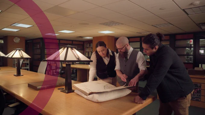Three people in the William Ready Division of Archives and Research Collections looking at a large book. A faded maroon half circle overlays the left side of the image.