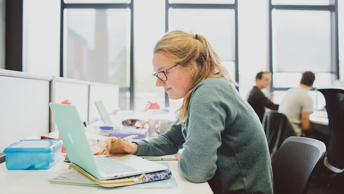 A student sits at a library desk with an open laptop in front of them.