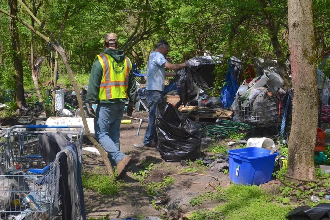 municipal workers gather homeless people's belongings in rash bags in an deserted encampment.