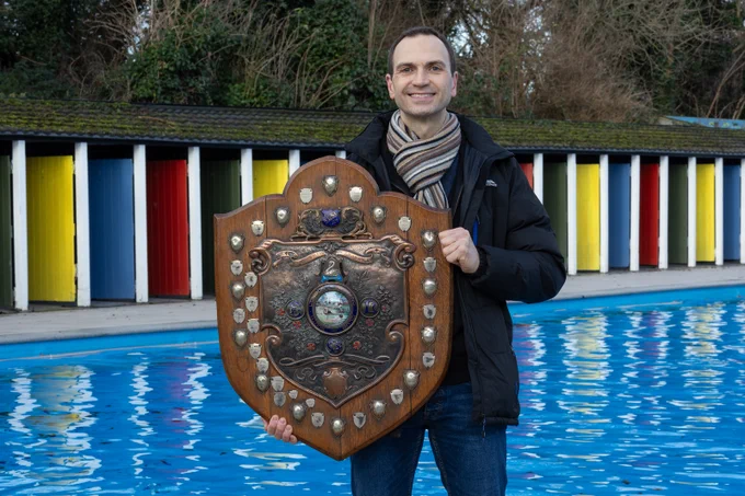 Man holding a prize shield stood in front of Tooting Bec Lido's outdoor swimming pool