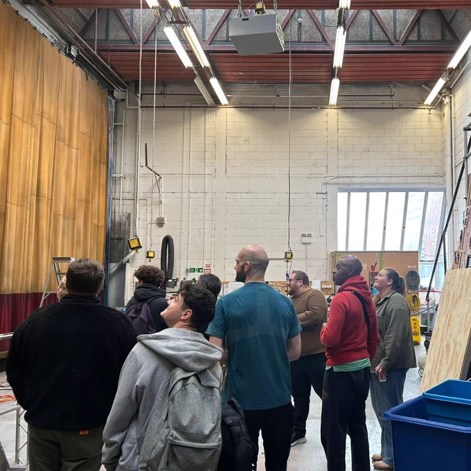 A group of people stands in a large backstage area, looking at a set with tall curtains and stage props, including large painted flats resembling a building façade. The space is industrial with exposed beams and bright lighting.