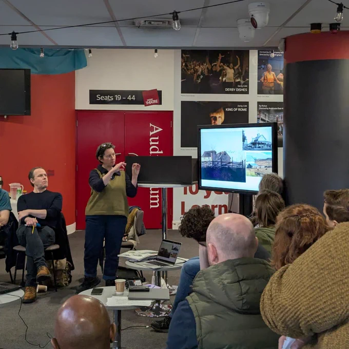 A presenter stands in front of an audience in a brightly lit room, pointing to a screen displaying scenic images. The audience is seated, with some people taking notes, while posters and signage, including "Seats 19-44," are visible in the background.