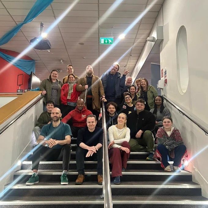 A group photo of around 20 people sitting on a staircase in a brightly lit indoor space. The group appears cheerful, with a mix of casual and winter attire, and blue fabric decorations hanging from the ceiling.